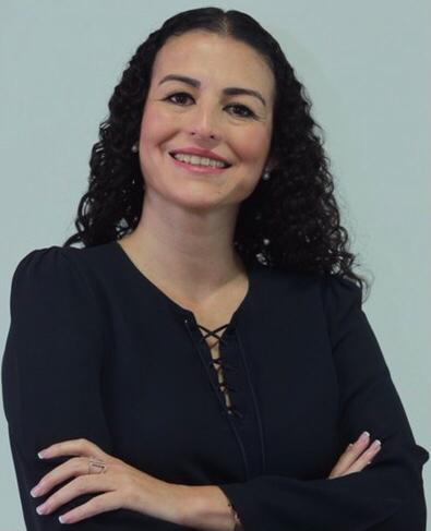 Mujer blanca con blusa negra y cabello largo crespo sonriendo a la cámara con un fondo blanco