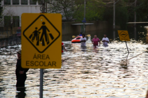 People wade through waist-deep floodwaters on a street, with school zone and traffic signs partially submerged. In the background, a small inflatable boat floats near more individuals navigating the flooded area.